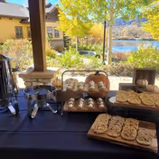 Outdoor table set with trays of assorted cookies and a beverage dispenser, with a scenic view of a pond, trees, and buildings in the background under a sunny sky.