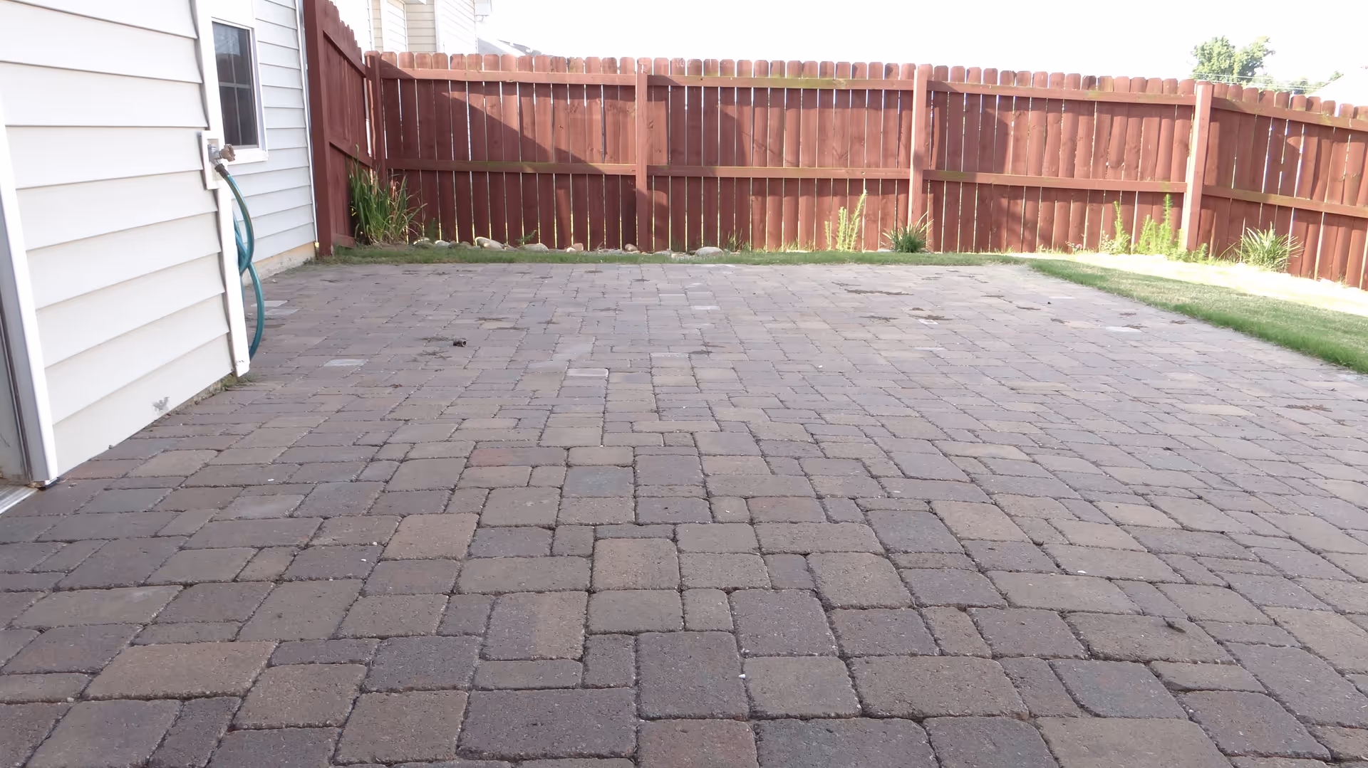 A paved outdoor patio area with interlocking stone tiles, adjacent to a white building wall with a garden hose attached. The patio is enclosed by a wooden fence with some grass and plants along the edges.