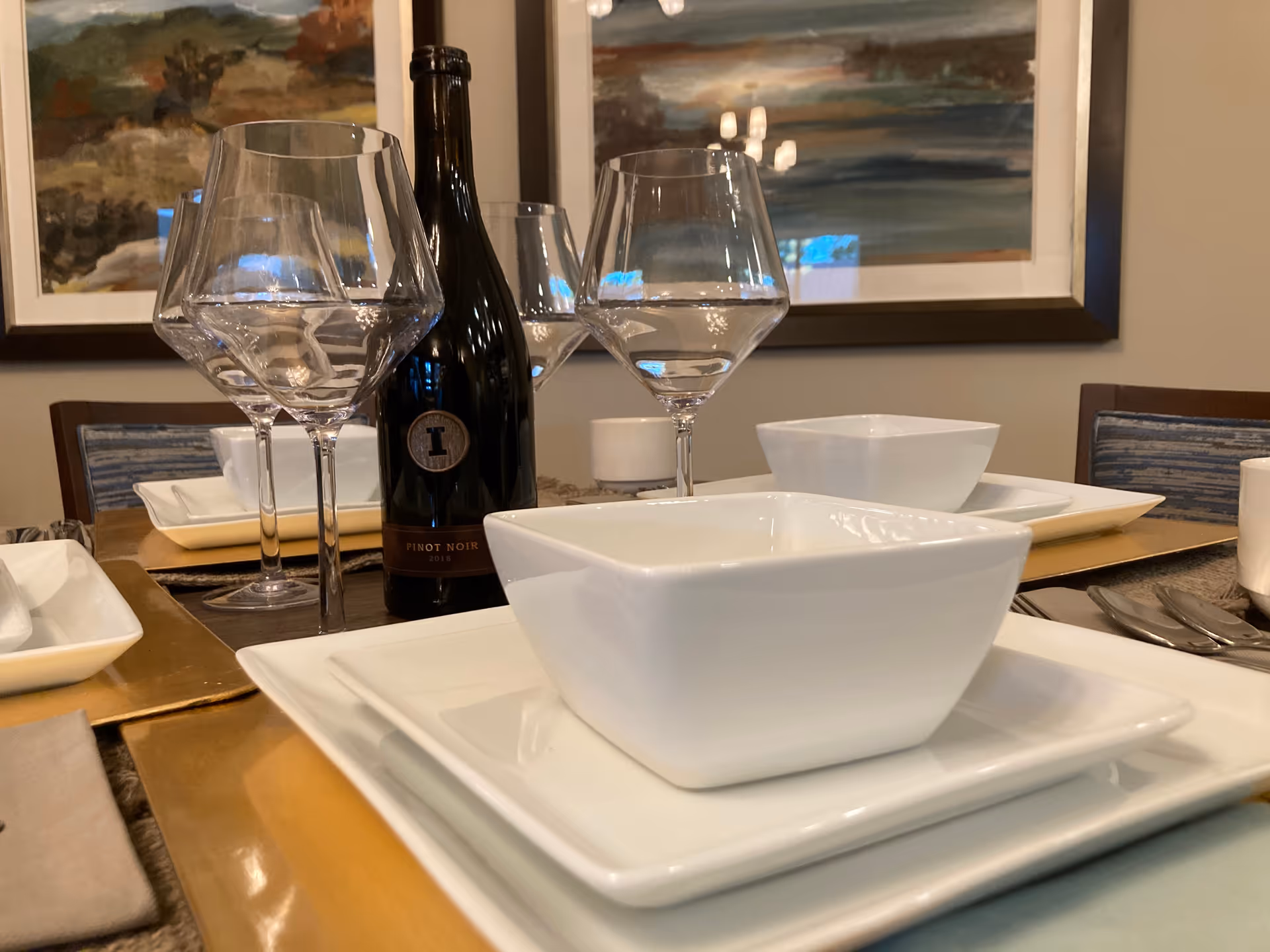 Close-up of a dining table set with white square plates, bowls, wine glasses, and a bottle of Pinot Noir wine. The background shows framed landscape paintings and upholstered chairs.