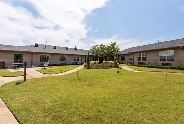 A sunny outdoor courtyard area with well-maintained green grass, a central landscaped garden with shrubs and a tree, surrounded by a single-story building with multiple windows and doors. There are paved walkways leading through the courtyard and a swing set visible on the left side.