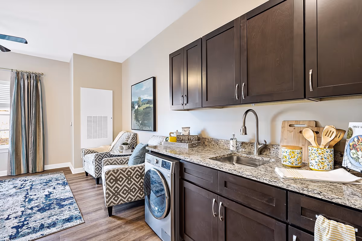 Interior view of a room featuring a kitchenette with dark wood cabinets, a granite countertop, a stainless steel sink, and a front-loading washing machine. Next to the kitchenette are two patterned armchairs, a small side table with a plant, a wall painting, and a window with striped curtains. The floor is wooden with a blue and white area rug.
