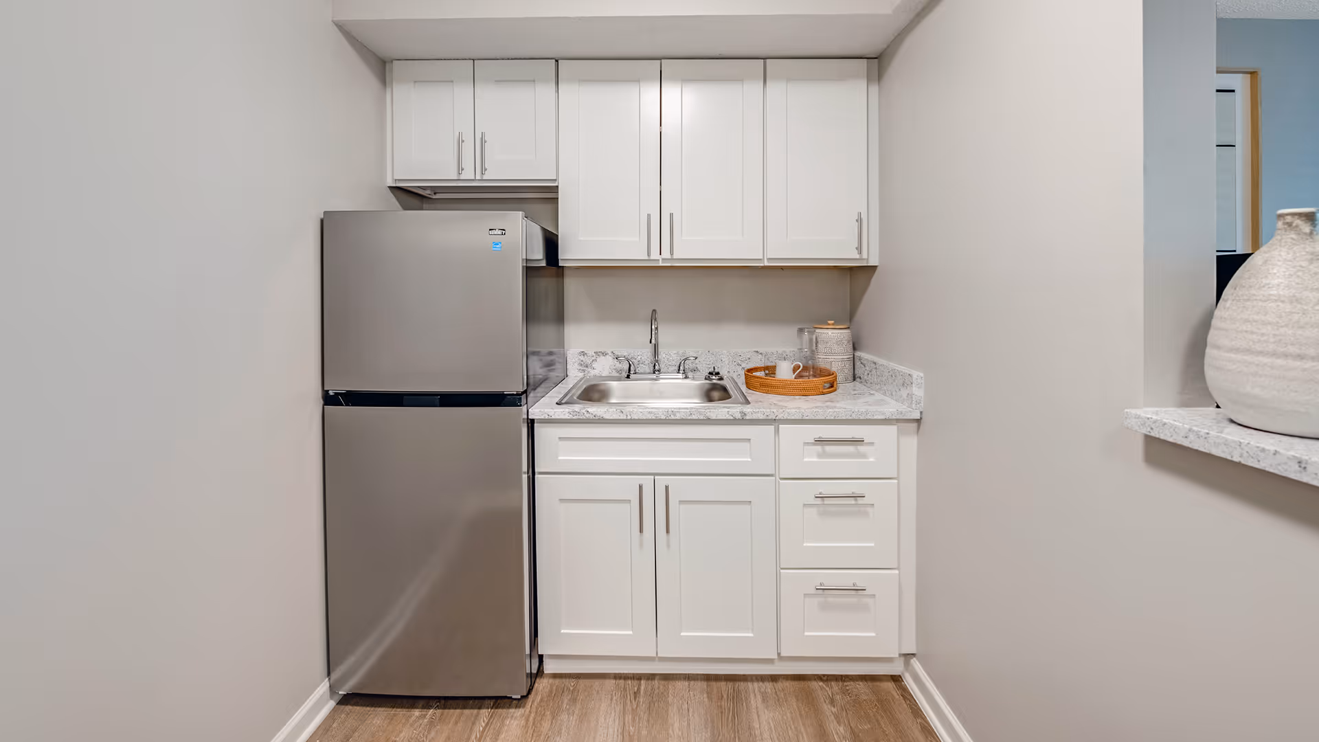 A small kitchen area featuring white cabinets, a stainless steel refrigerator, a stainless steel sink with a faucet, and a countertop with a decorative tray and container. The walls are painted light gray and the floor has wood-style flooring.