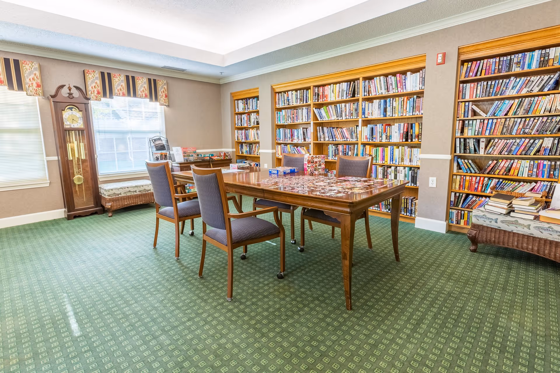 A bright room with green patterned carpet, a wooden table with four chairs, and several bookshelves filled with books along the walls. There is a grandfather clock near two windows with valances, and a cushioned bench with books stacked on it.