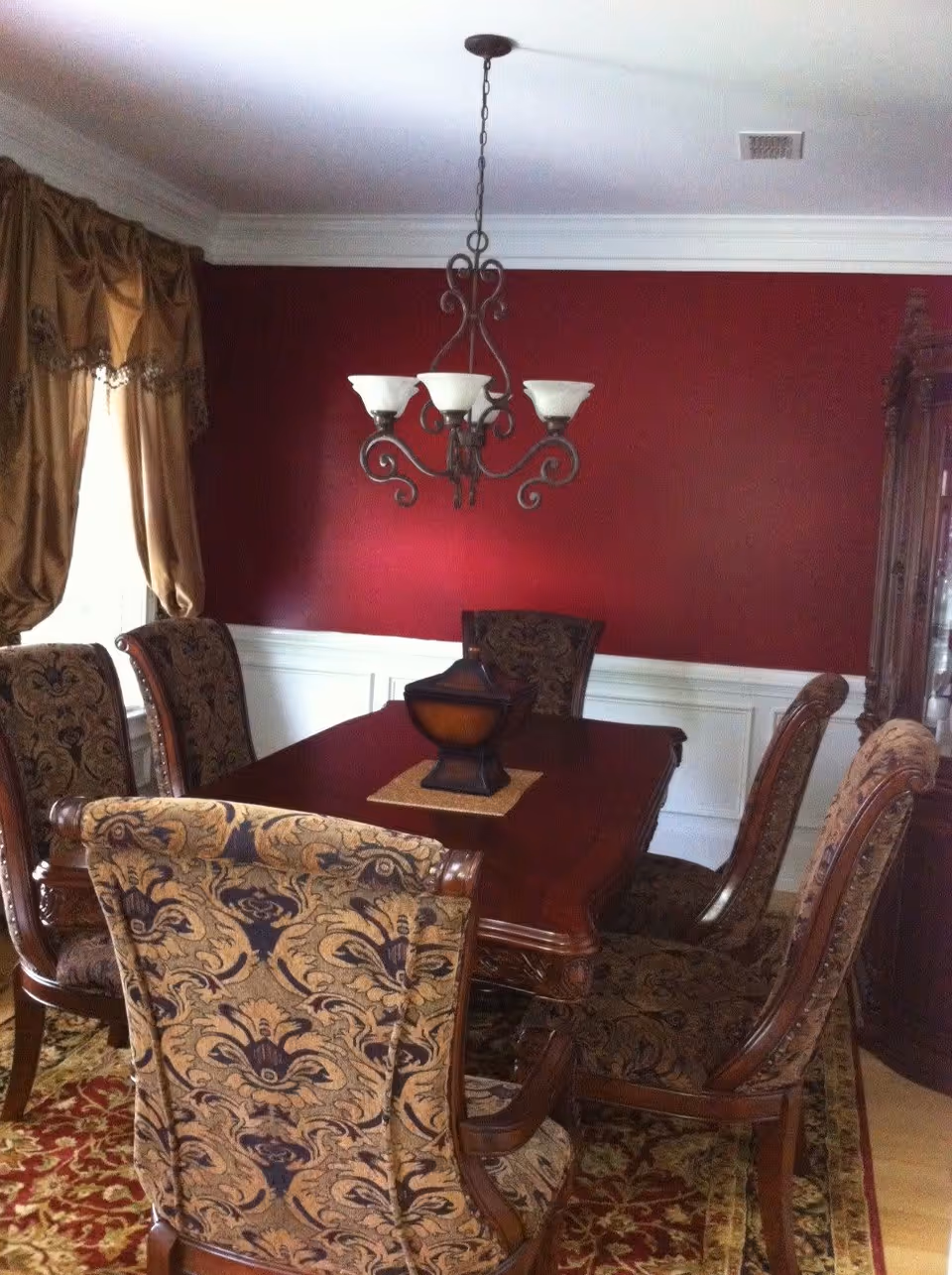 A formal dining room with a dark wooden table surrounded by six upholstered chairs with intricate patterns. The walls are painted deep red with white wainscoting, and there is a decorative chandelier hanging above the table. A window with gold curtains is on the left side, and a wooden cabinet is partially visible on the right.