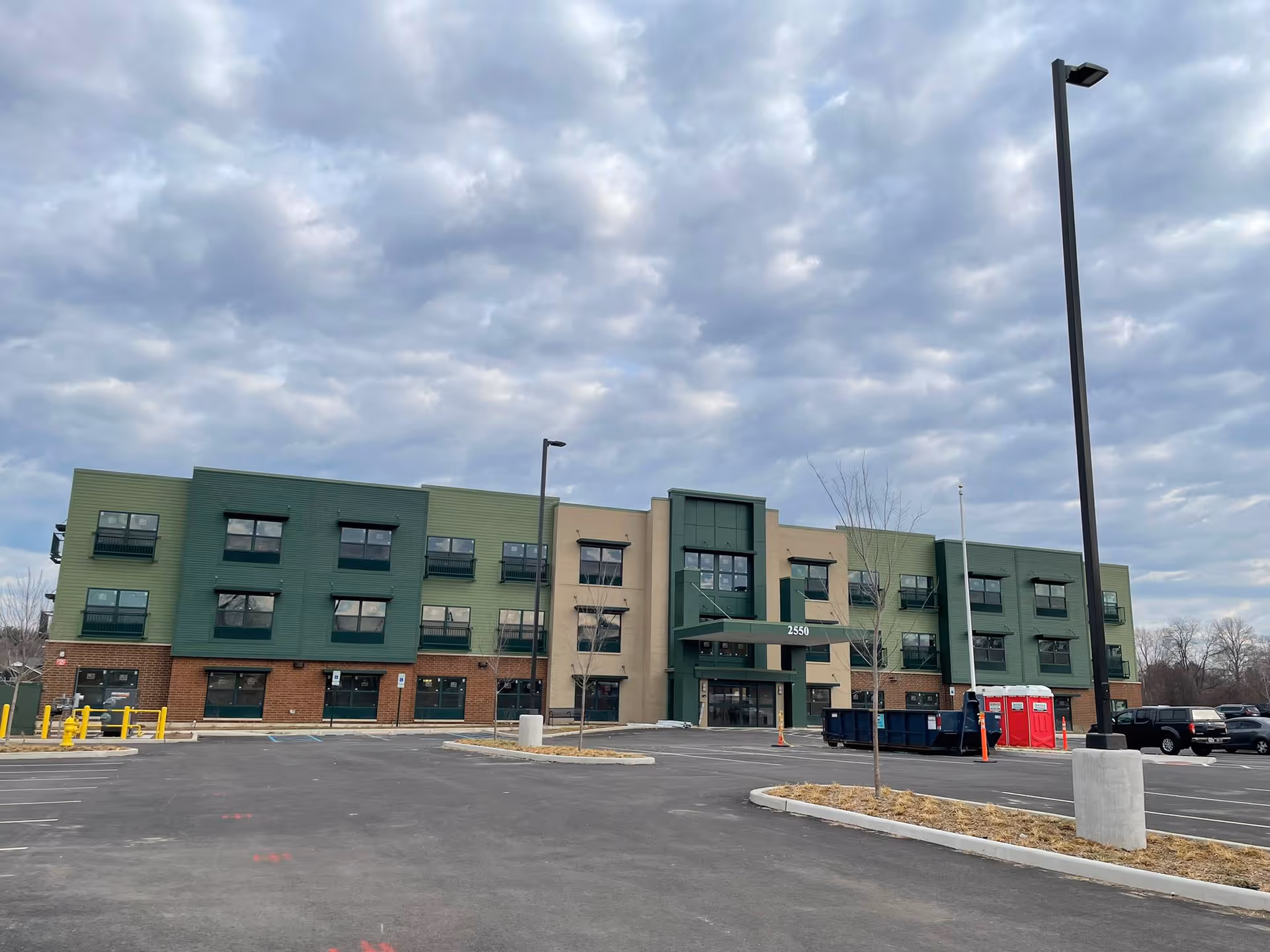 Exterior view of a three-story building with green and beige walls and multiple windows, situated in a large parking lot under a cloudy sky. The building has the number 2550 above the entrance, with some construction equipment and portable toilets visible on the right side of the parking lot.