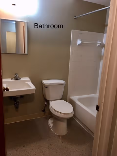 A small bathroom with a white toilet, a white sink with a silver faucet, a mirror above the sink, and a bathtub with white tiled walls and a shower curtain rod.