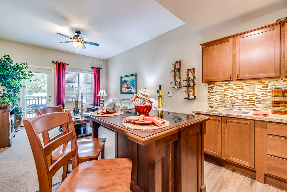 Open-plan kitchen and living area with a wooden island and bar stools, wood cabinets, and a seating area by windows with red curtains.