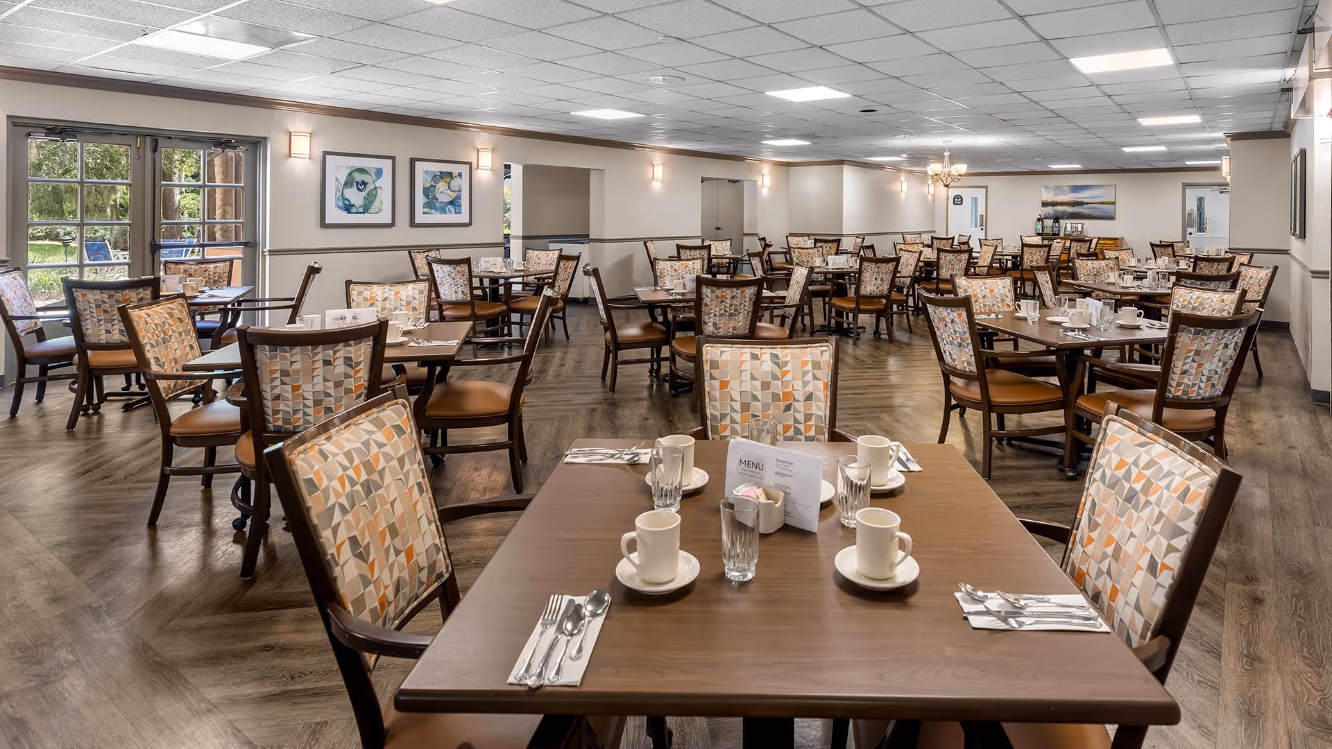 A spacious dining room with multiple wooden tables and chairs arranged neatly. Each table is set with white cups, glasses, silverware, and a menu card. The room has large windows on one side letting in natural light and decorated walls with framed artwork. The floor is wooden, and the ceiling has recessed lighting.