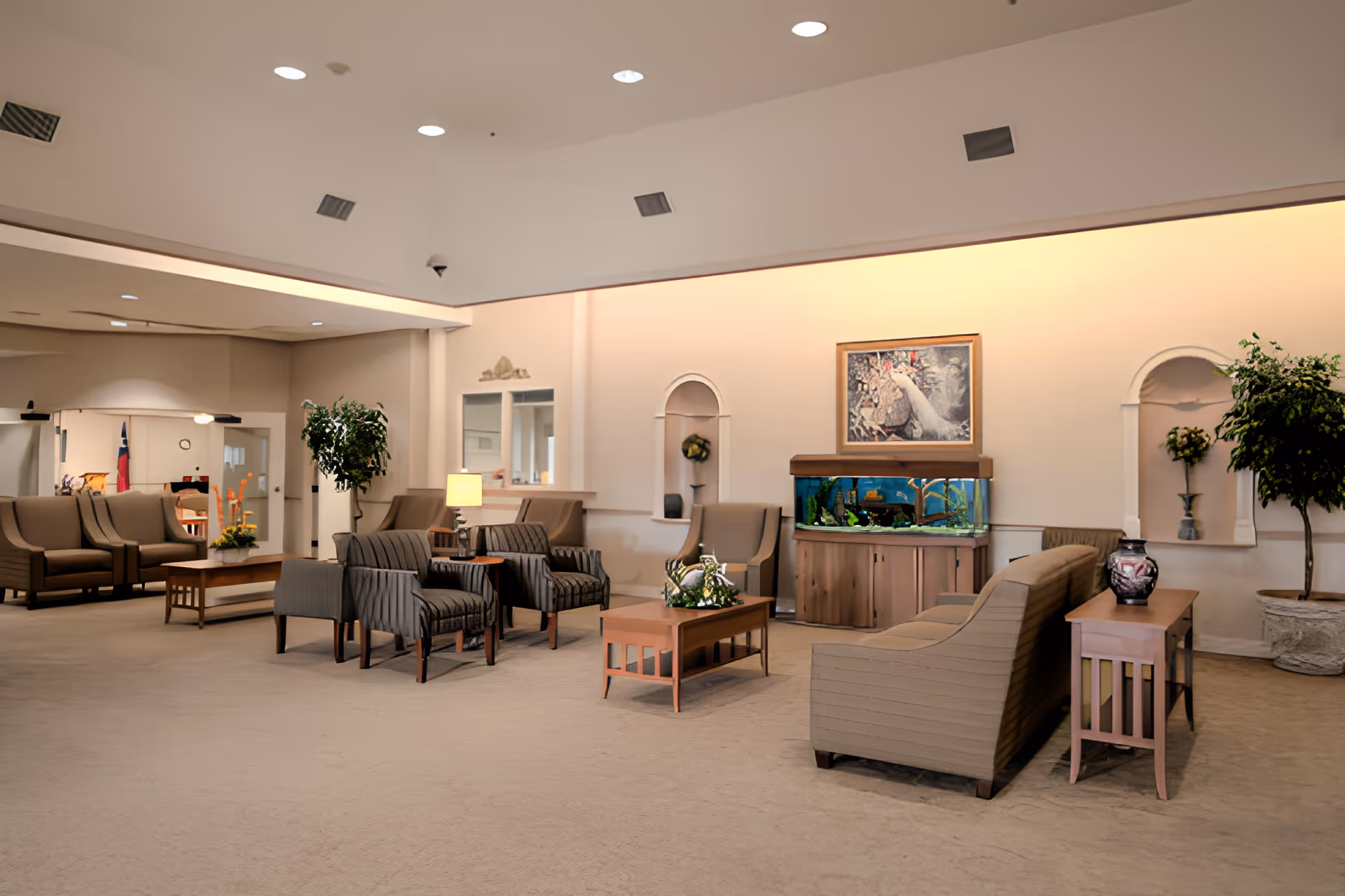 Open senior living facility lounge with sofas, armchairs, coffee tables, an aquarium, and potted plants.