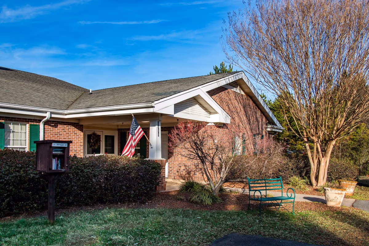 Exterior view of a single-story brick building with green shutters and a covered porch displaying an American flag. There is a small green bench and a little free library box in the front yard, surrounded by bushes and trees under a clear blue sky.