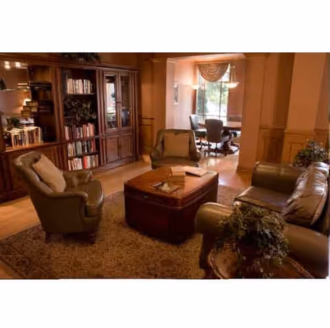 Cozy communal living room with leather sofas and armchairs arranged around a central ottoman, a bookcase along the wall, and a dining area visible through an adjacent opening.