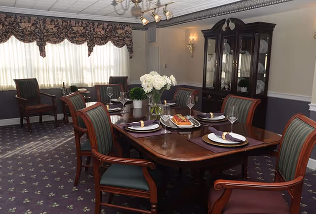 A formal dining room with a polished wooden dining table set for six people. Each place setting has a white plate, a folded napkin with a napkin ring, and a wine glass. A vase with white flowers and a small potted plant are placed in the center of the table. The room has carpeted flooring with a patterned design, large windows with floral valances, and a dark wooden china cabinet against the wall.