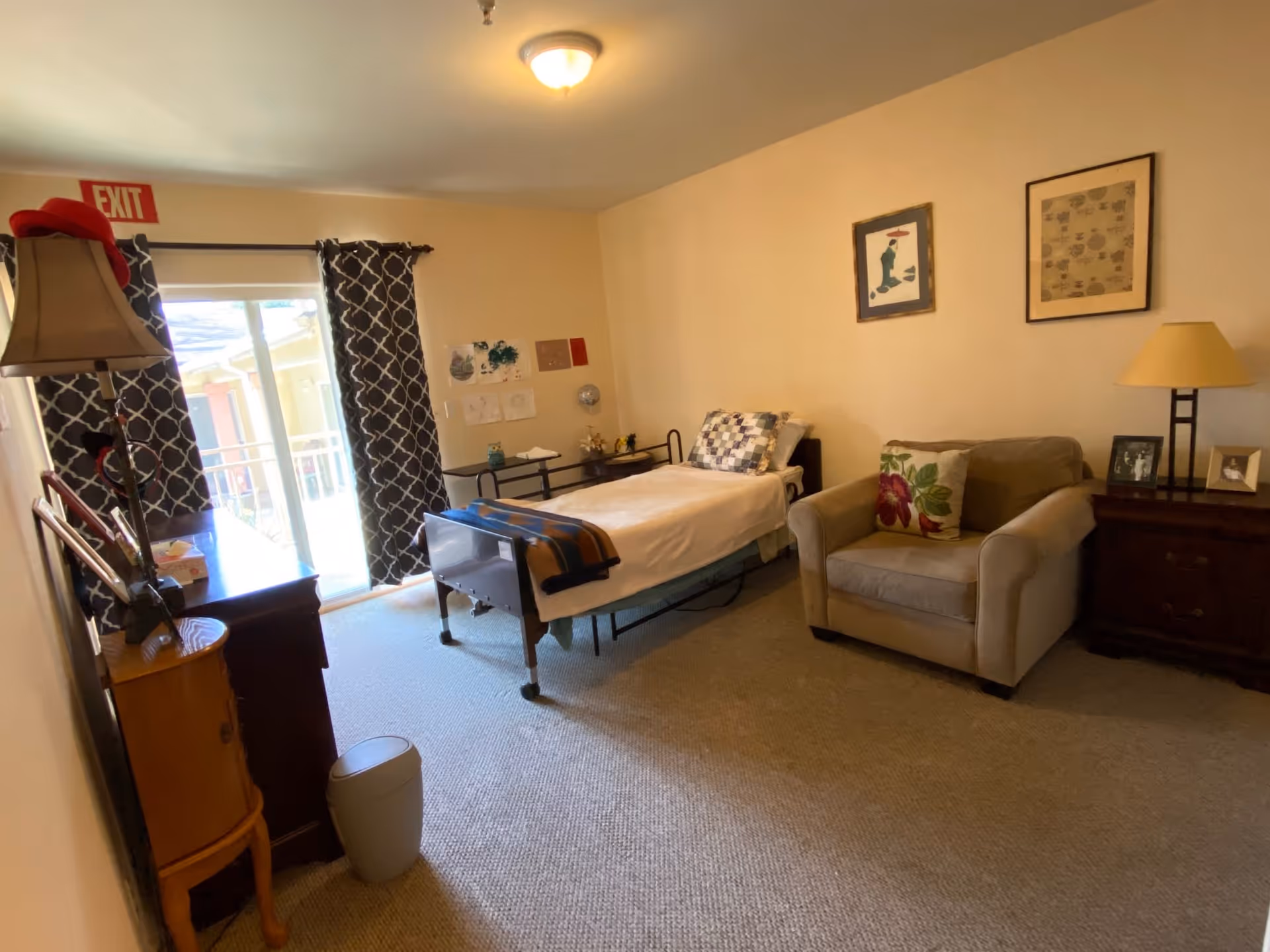 A cozy senior living bedroom with a single bed covered with a white blanket and a blue and brown striped throw. There is a beige armchair with a floral cushion next to a wooden nightstand with a lamp and framed photos. The room has beige walls with two framed pictures hanging above the armchair. A dresser with a lamp and other items is near a sliding glass door with patterned curtains, letting in natural light. An exit sign is visible above the door.