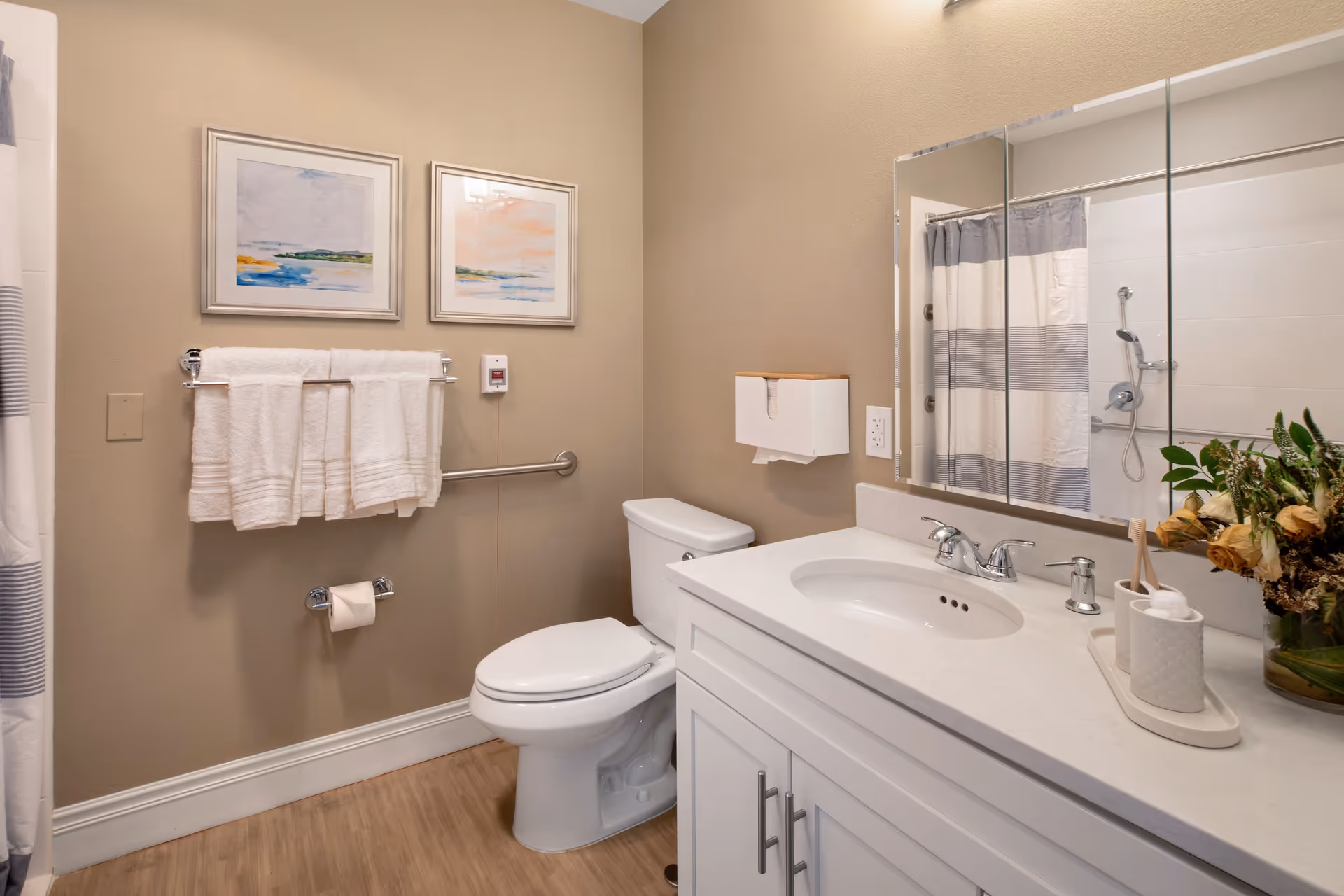 A clean and well-lit bathroom featuring a white toilet, a white vanity with a sink and chrome faucet, a large mirror above the sink, and a shower with a striped curtain. The walls are beige, decorated with two framed landscape paintings. There are white towels hanging on a chrome towel rack, a toilet paper holder, a wall-mounted tissue dispenser, and a grab bar near the toilet. A vase with flowers and bathroom accessories are placed on the vanity countertop.