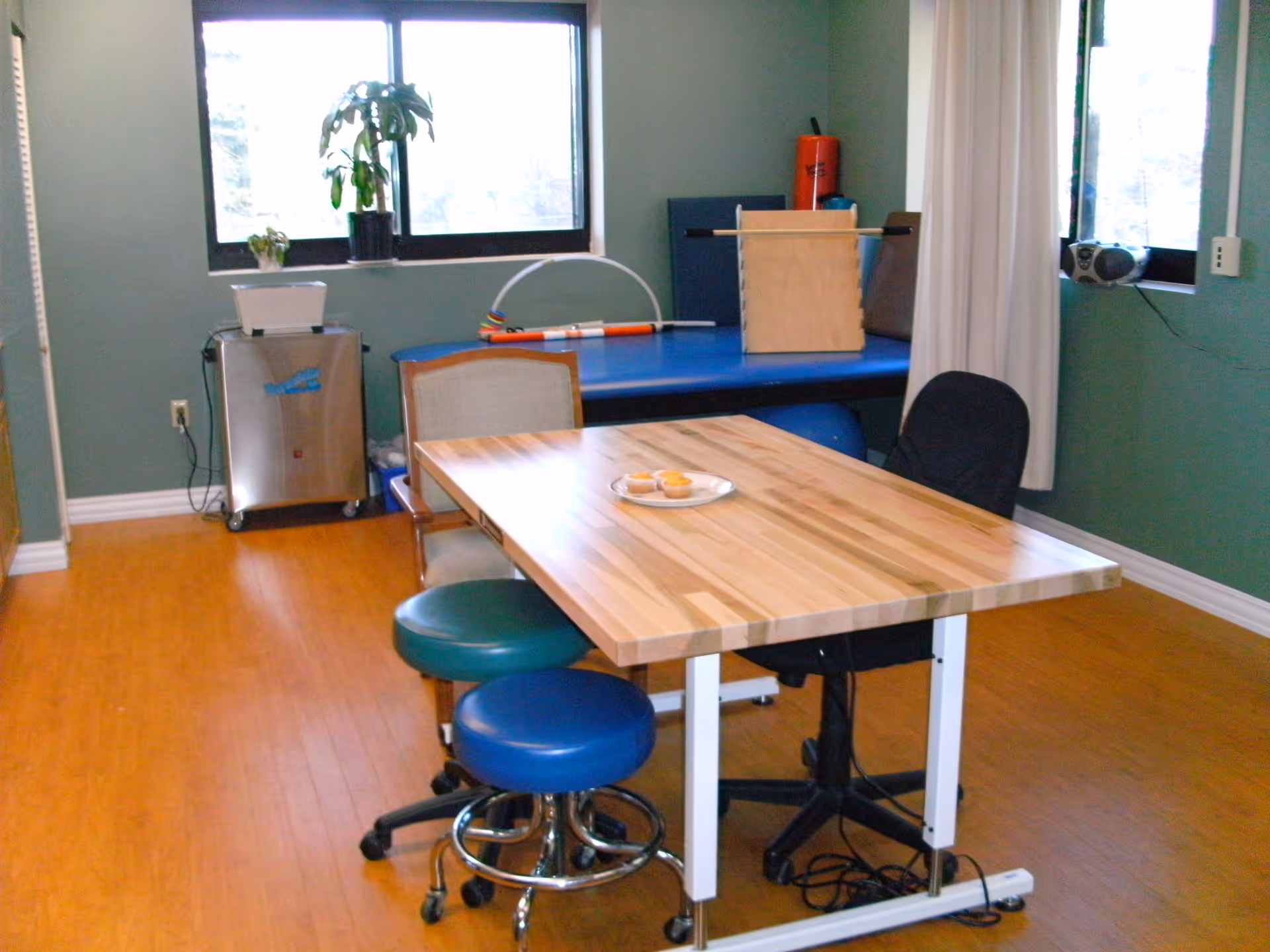 A room with a wooden table surrounded by various chairs including a black office chair and two round stools. On the table is a plate with some muffins. In the background, there is a blue therapy or massage table with exercise equipment and a plant on a small metal cabinet near a window. The room has green walls and wooden flooring.