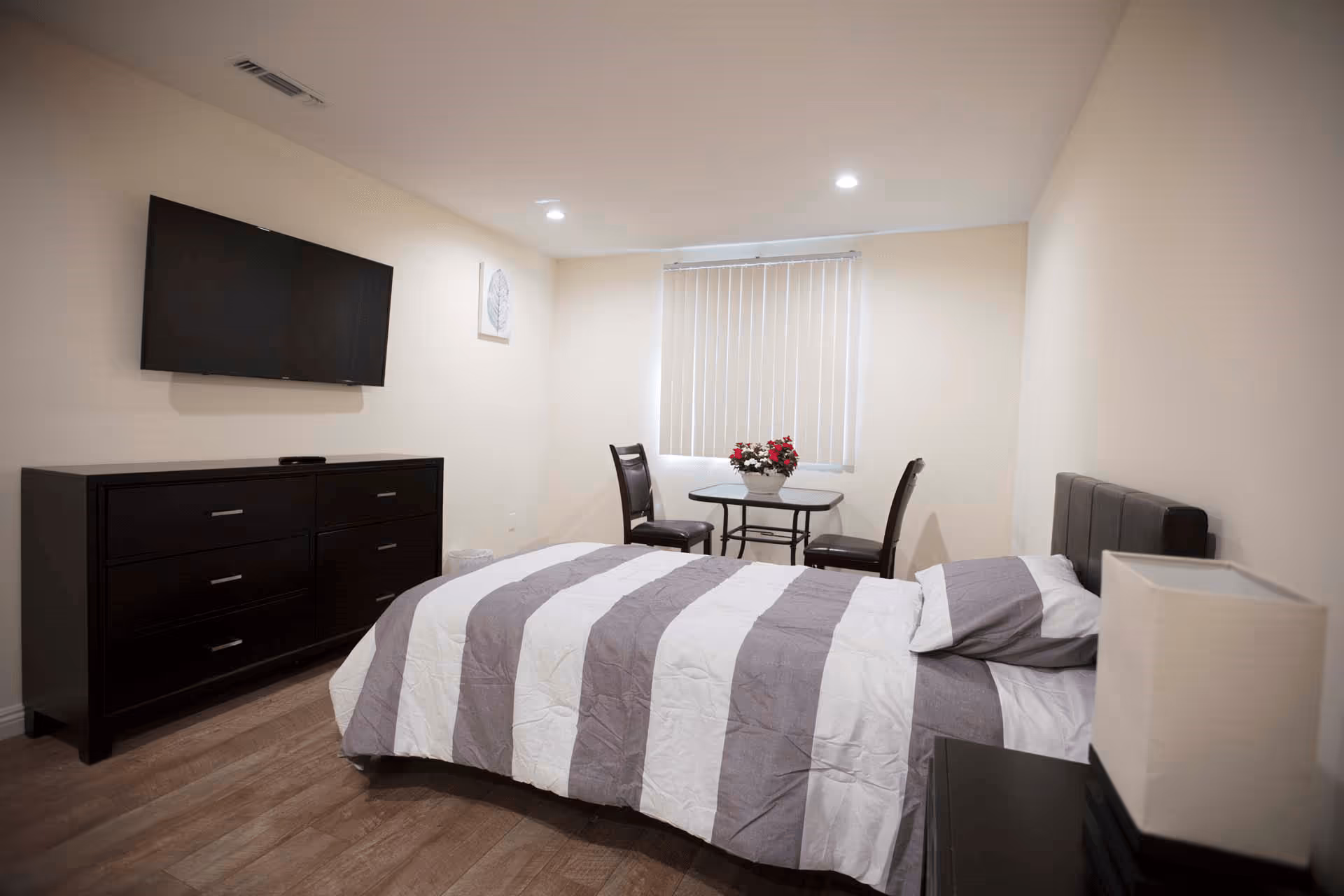 A bedroom with a bed covered in a gray and white striped comforter, a dark wooden dresser with a flat-screen TV mounted above it, a small table with two chairs and a flower arrangement near a window with vertical blinds, and a nightstand with a lamp.