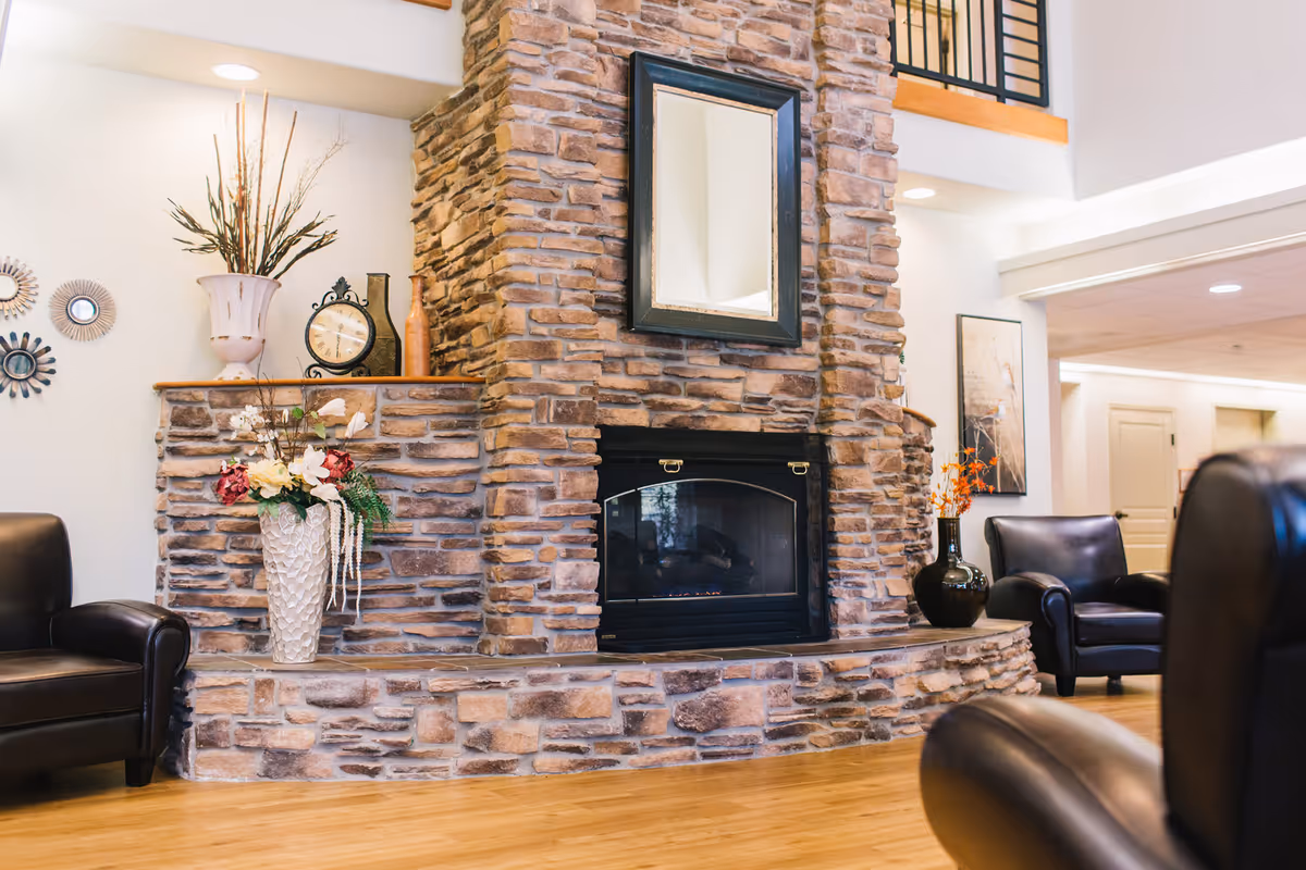 A cozy living room area featuring a large stone fireplace with a black-framed mirror above it. The fireplace mantel is decorated with vases, a clock, and floral arrangements. Surrounding the fireplace are several black leather armchairs on a wooden floor, with light-colored walls and a high ceiling.