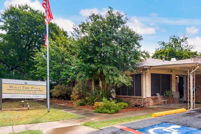 Front entrance of Peach Tree Place nursing and Alzheimer's care with a sign, flagpole, covered entry, and surrounding trees.