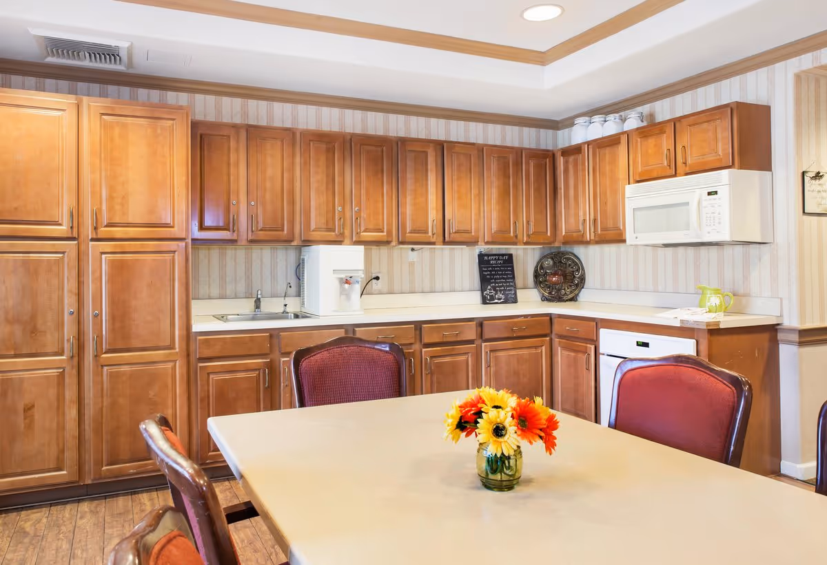 Community kitchen with wooden cabinets, microwave, sink and a dining table topped with a vase of flowers.