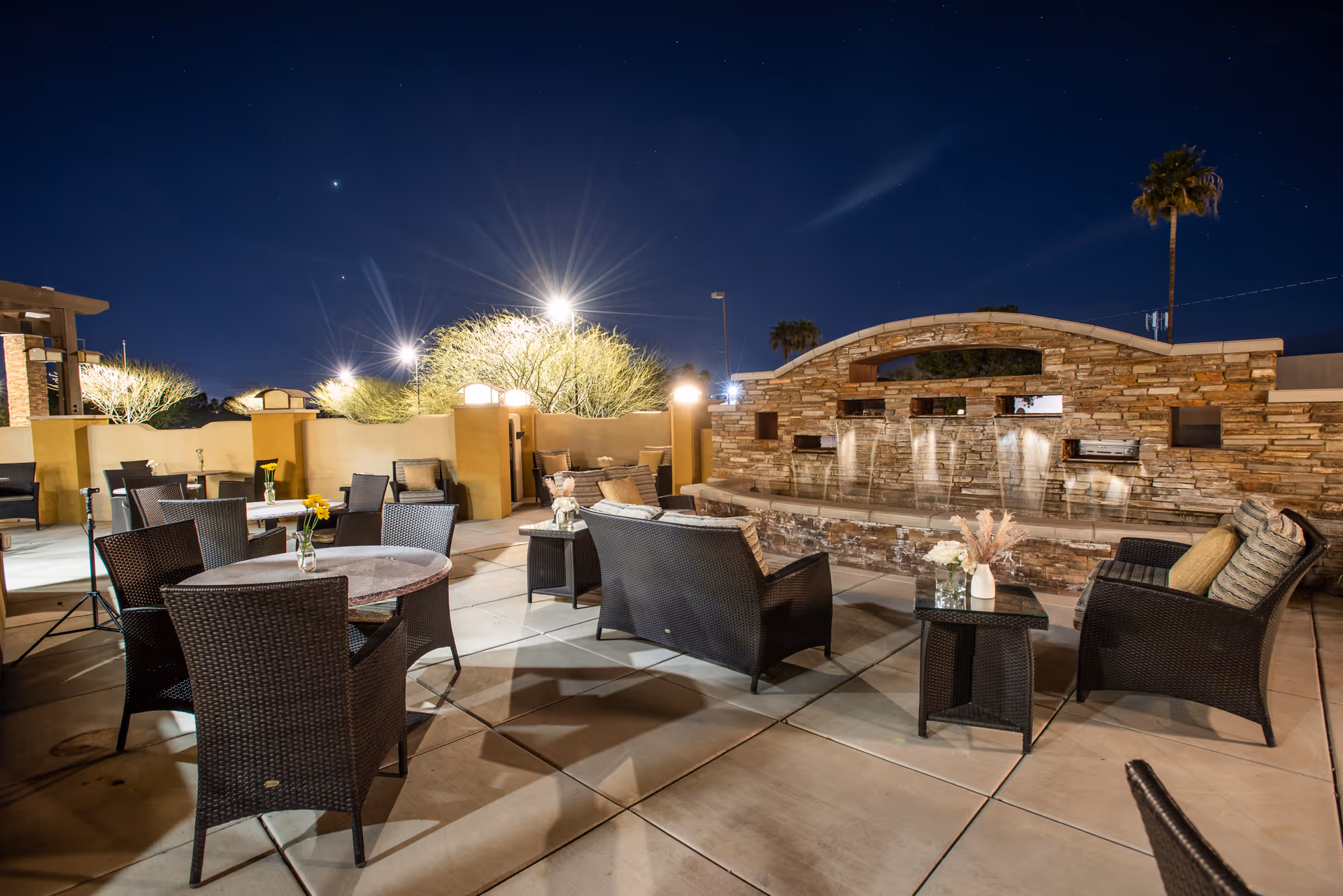 Outdoor patio area at night with multiple wicker chairs and tables arranged around a stone water feature with cascading water. The area is well-lit with several bright lights and has decorative plants and flowers on the tables. A palm tree is visible in the background under a clear night sky.