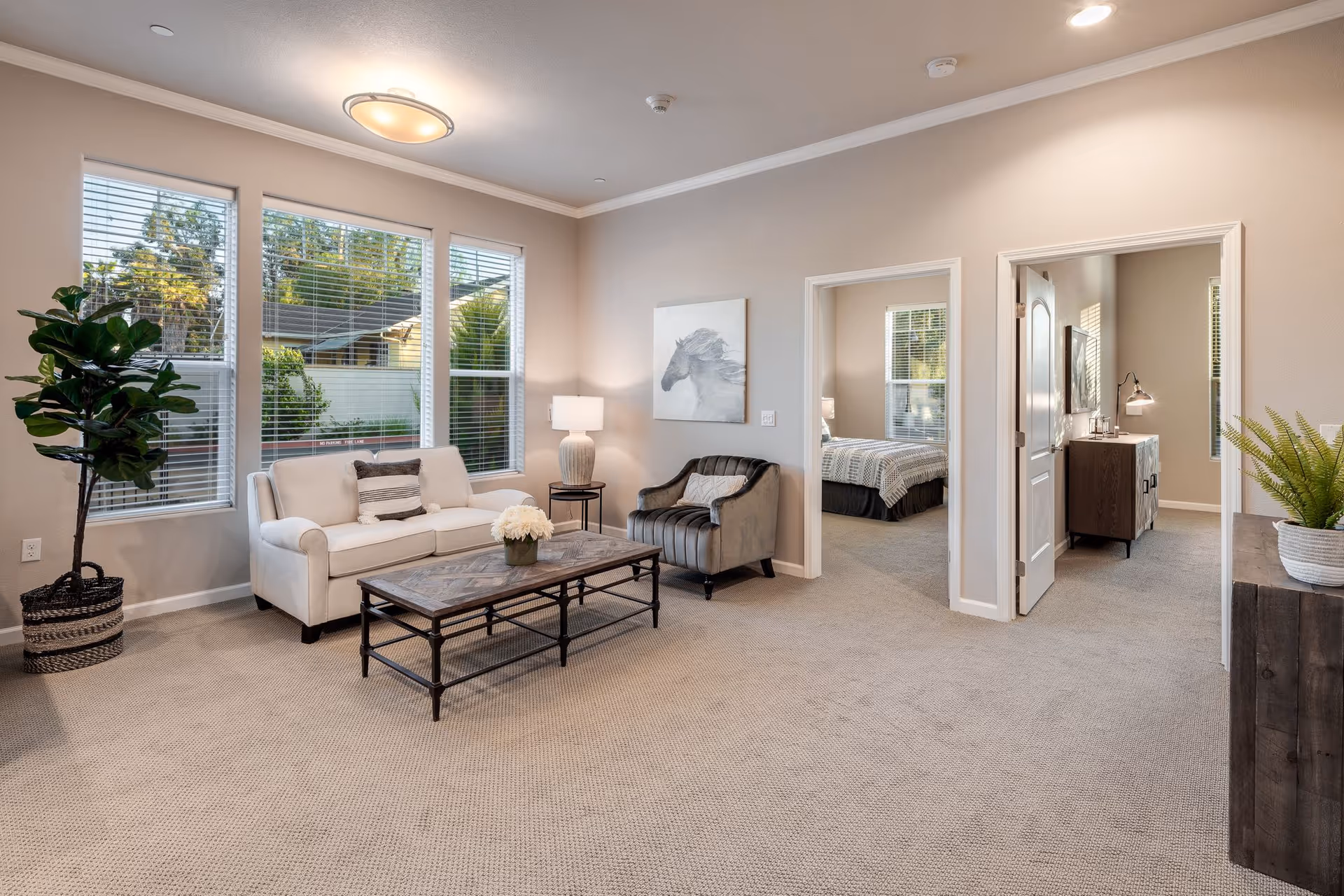 A bright living room with beige carpet, a white sofa with a striped pillow, a gray armchair, a wooden coffee table with a flower pot, a floor lamp, and large windows showing greenery outside. Two doorways lead to a bedroom and another room with a dresser and lamp.