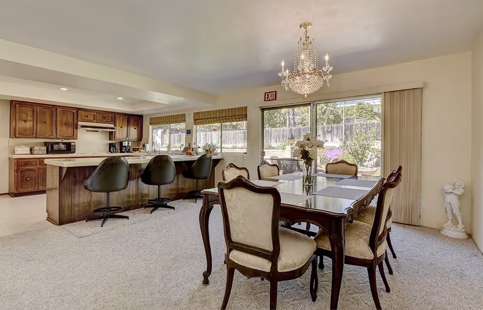 A spacious dining area with a wooden dining table and six upholstered chairs. A crystal chandelier hangs above the table. To the left, there is a kitchen with wooden cabinets, a countertop with three black swivel bar stools, and various kitchen appliances. Large windows and a sliding glass door provide natural light and a view of the outdoor garden area. A small white statue is placed in the corner near the sliding door.