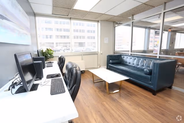A bright office lounge area with two black leather chairs at a white desk with two computer monitors and keyboards. Across from the desk is a dark blue tufted leather sofa and a white marble coffee table with gold legs. Large windows with blinds allow natural light to fill the room, and a glass partition separates this space from an adjacent room with a wooden table and chairs.
