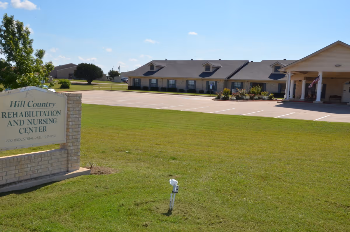 Exterior view of Hill Country Rehabilitation and Nursing Center showing a large green lawn, a parking lot with marked spaces, and a single-story building with a covered entrance and an American flag.