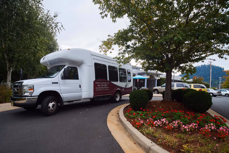 A white and maroon shuttle bus parked on a driveway near a landscaped area with a tree and colorful flowers. Several cars are parked in the background near a building entrance, under a cloudy sky.