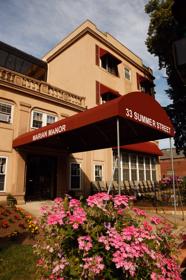 Entrance of Marian Manor building with a red awning displaying the address 33 Summer Street. The building is beige with multiple windows and red shutters. There are pink flowers and a garden area in the foreground, along with a row of chairs near the entrance.