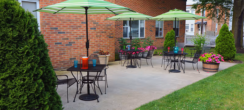 Outdoor patio area with three black metal tables, each shaded by a green and white striped umbrella. Each table has chairs around it and colorful drinkware on top. The patio is adjacent to a brick building with windows and surrounded by green grass, bushes, and flower pots with pink flowers.