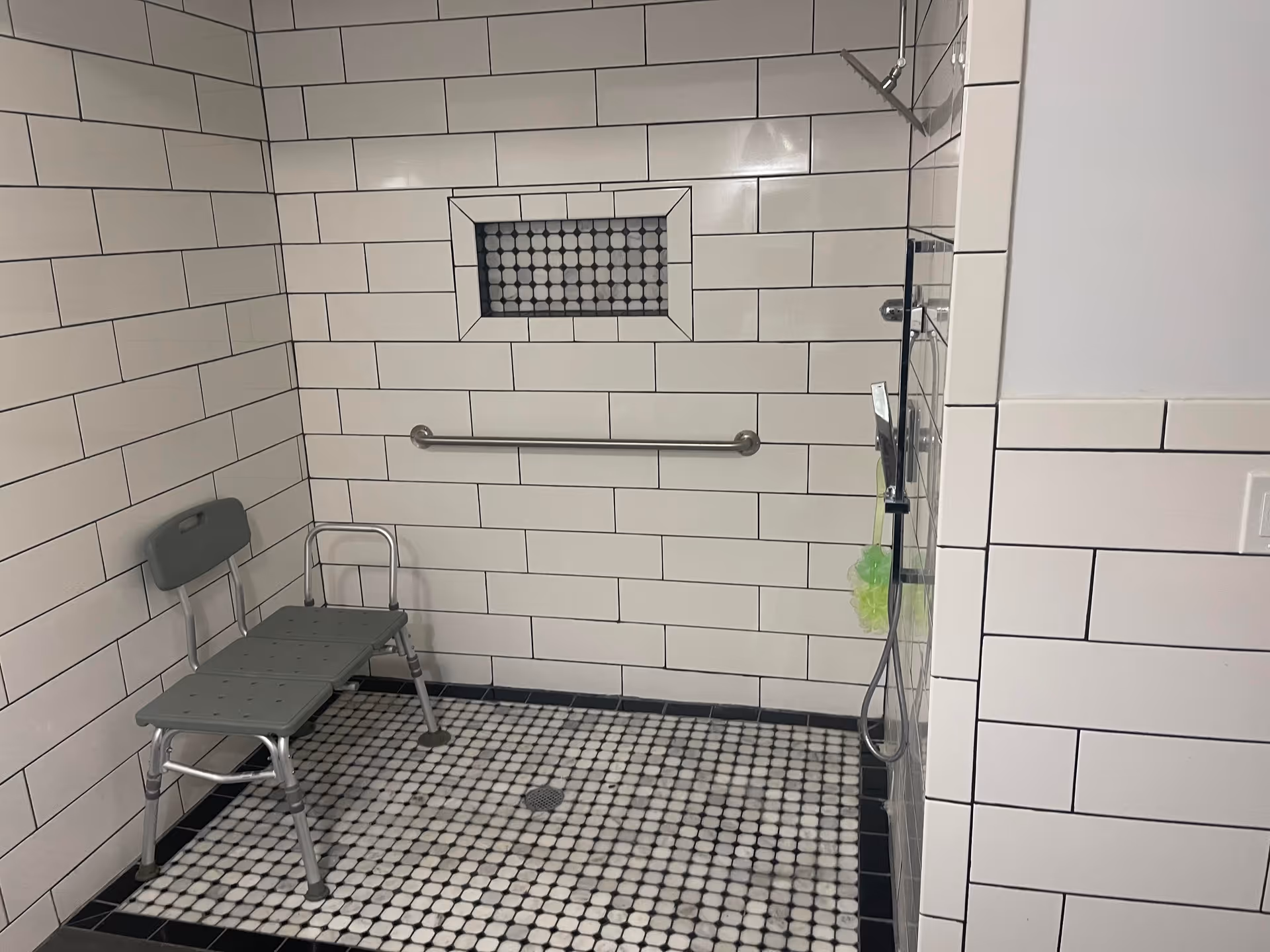 Accessible tiled shower with a gray shower chair, horizontal grab bar, handheld shower head, and mosaic floor tiles.
