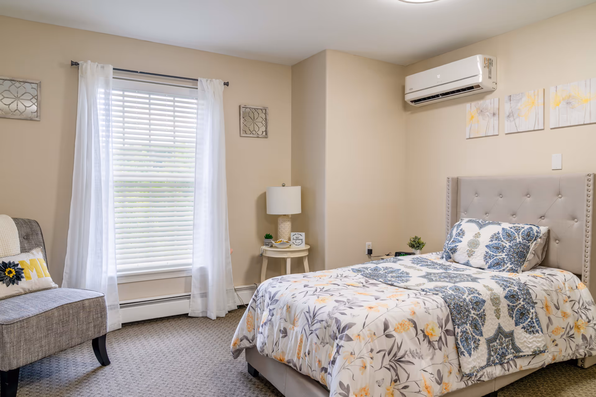 A cozy bedroom with a single bed featuring a floral patterned comforter and matching pillow. There is a beige upholstered headboard with button tufting. A small round side table with a lamp and decorative items sits next to the bed. A gray upholstered chair with a decorative pillow is positioned near a window with white blinds and sheer white curtains. The walls are painted beige and there is a white air conditioning unit mounted above the bed. Three floral paintings hang on the wall above the headboard.