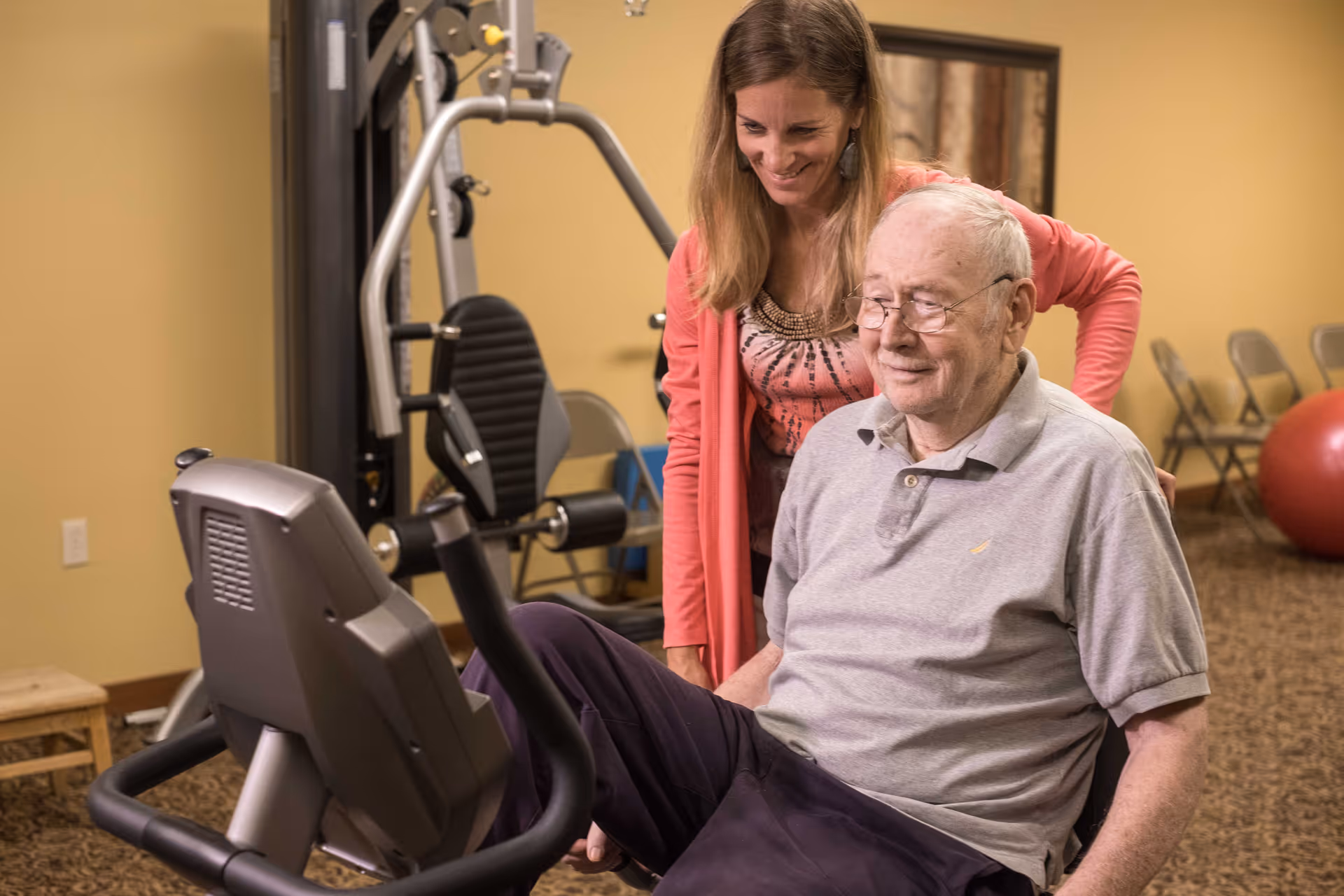 An elderly man using a seated exercise machine in a fitness room, assisted by a smiling woman standing beside him. The room has exercise equipment, folding chairs, and a large red exercise ball in the background.