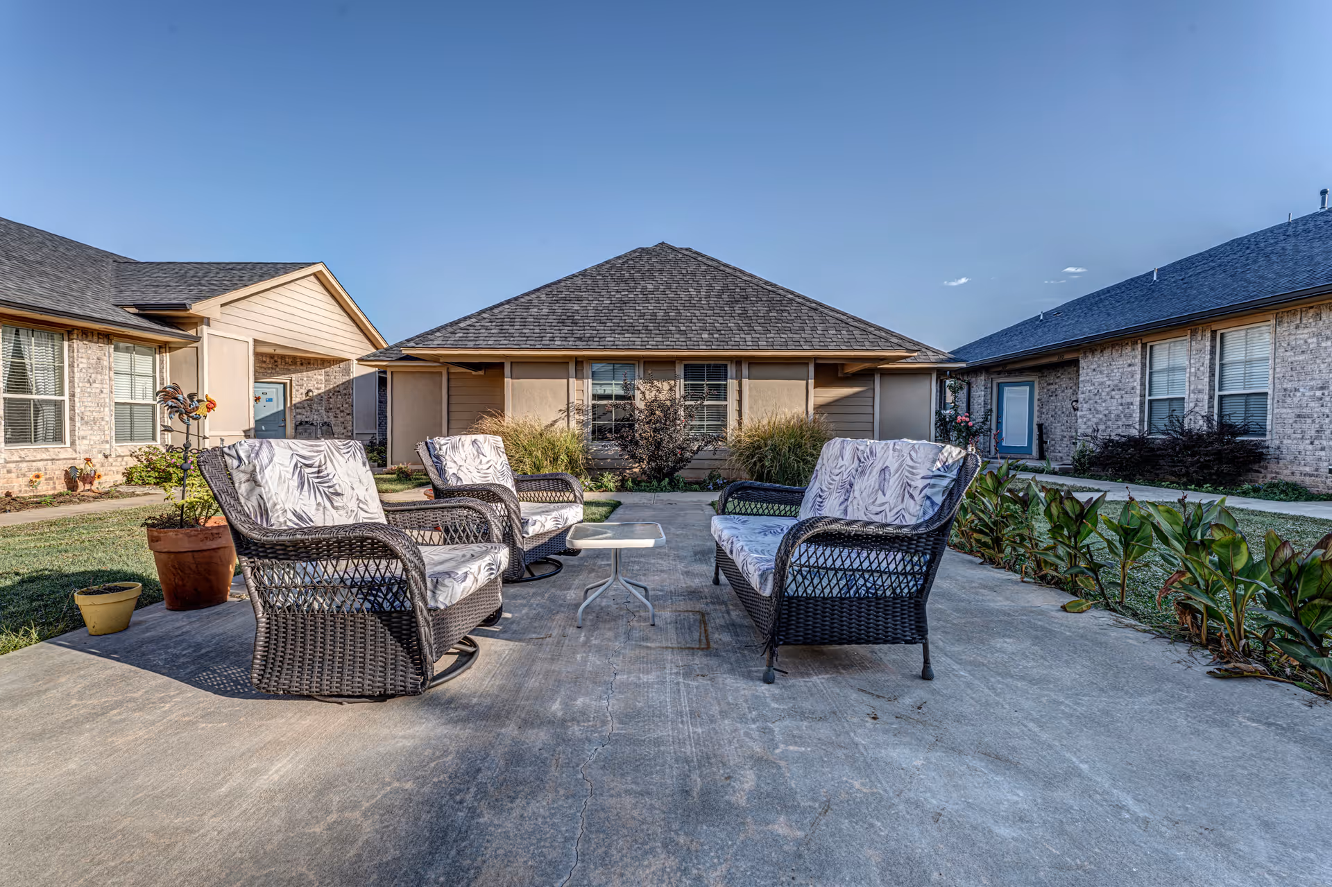 Outdoor patio area with four cushioned wicker chairs arranged around a small white table on a concrete surface, surrounded by low plants and residential buildings under a clear blue sky.