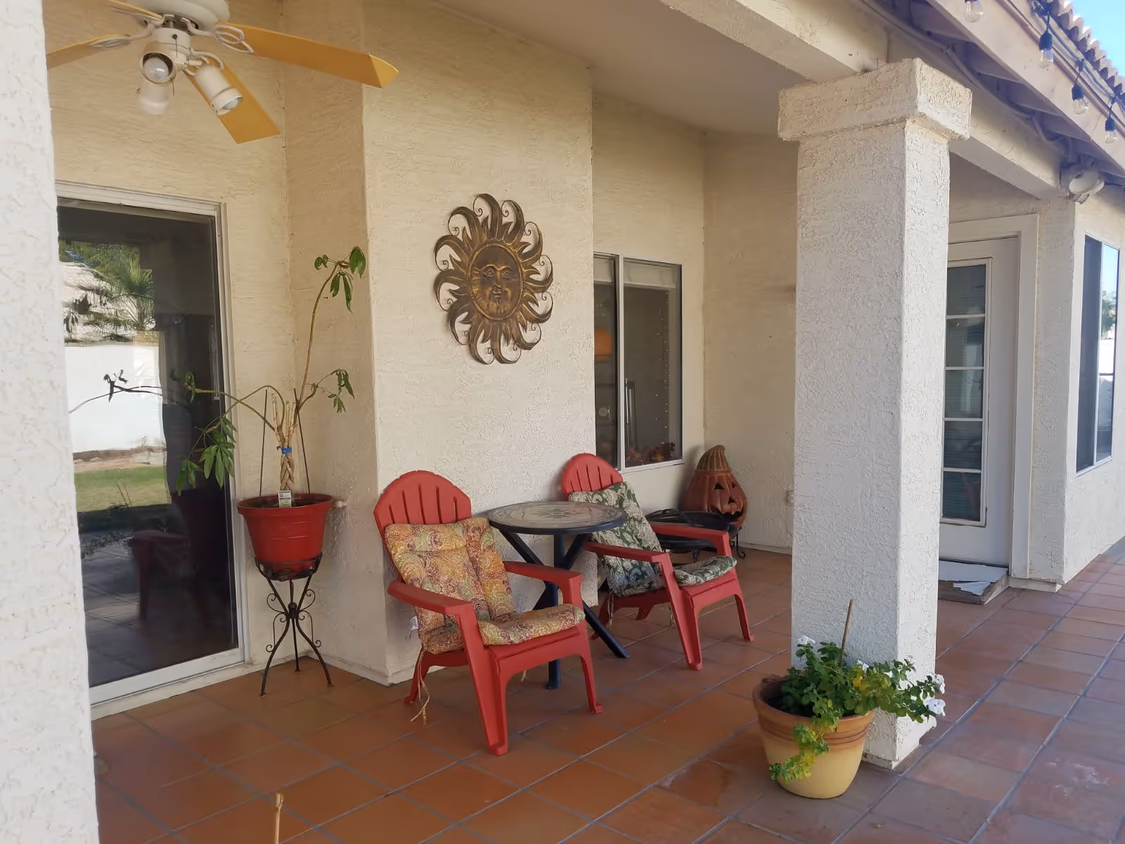 Outdoor covered patio area with two red chairs with cushions, a small round table between them, a potted plant on a stand, a decorative sun wall hanging, and a potted plant on the tiled floor near a white column.