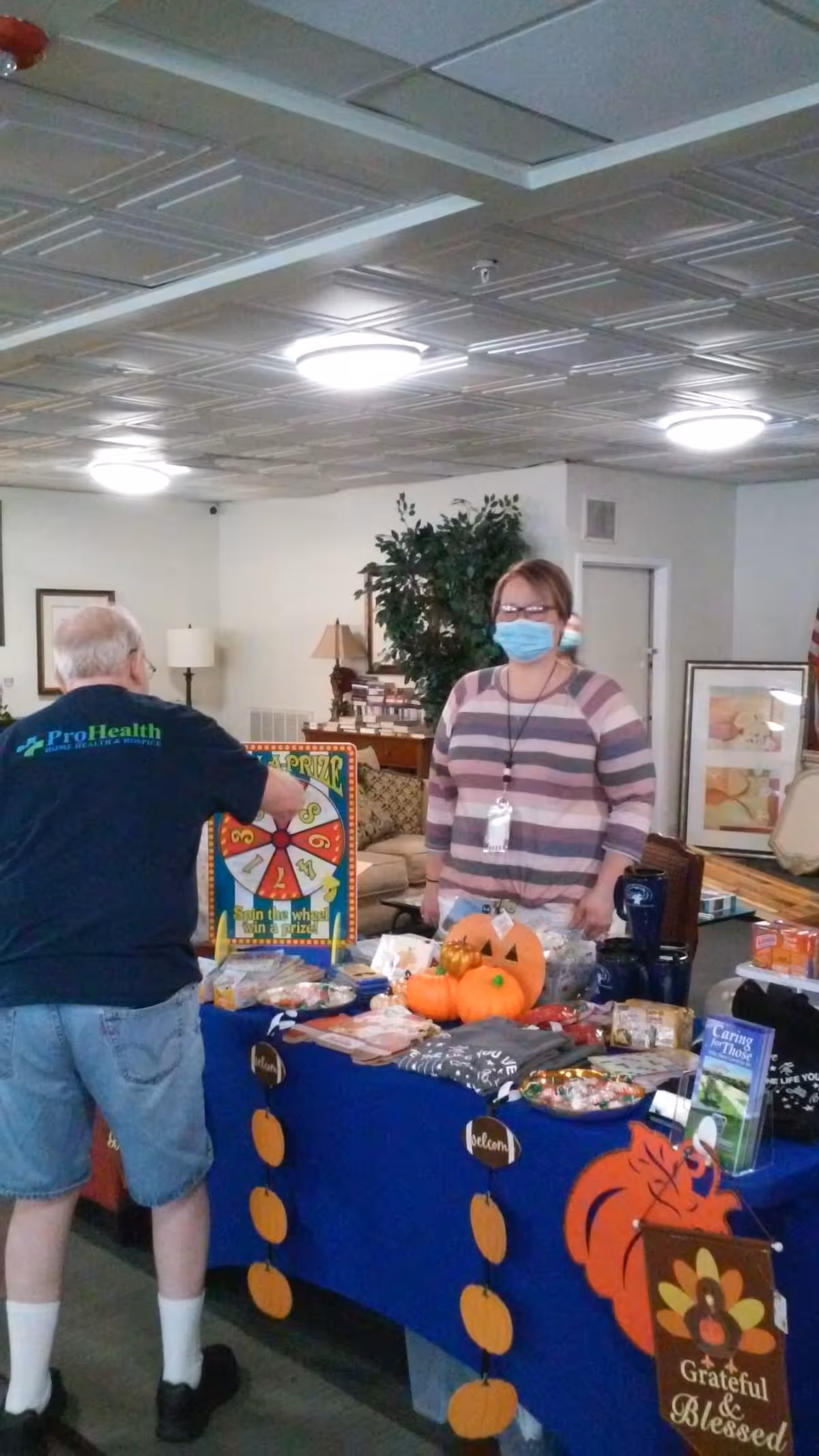 An indoor community room with a man spinning a prize wheel at a decorated table while a woman wearing a face mask stands behind the table. The table is covered with a blue cloth and decorated with small pumpkins and autumn-themed items, including a sign that says 'Grateful & Blessed'. The room has a cozy atmosphere with a couch, lamp, and framed pictures in the background.