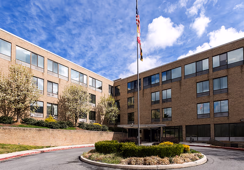 Exterior view of a three-story brick building with many windows, a circular driveway with a landscaped island in the center, and a tall flagpole flying the American flag and other flags under a partly cloudy blue sky.