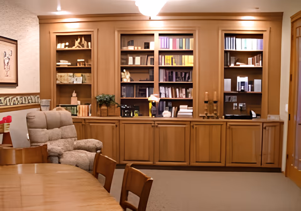 A cozy room with wooden bookshelves filled with books and decorative items, a comfortable beige recliner chair, a wooden table with chairs, and warm lighting creating a welcoming atmosphere.