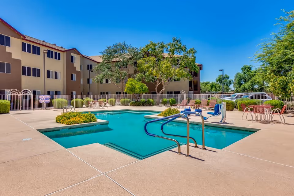 Outdoor swimming pool area at Discovery Point Retirement Community with clear blue water, surrounded by a concrete deck, poolside chairs, tables with umbrellas, and greenery. The multi-story residential building is visible in the background under a clear blue sky.