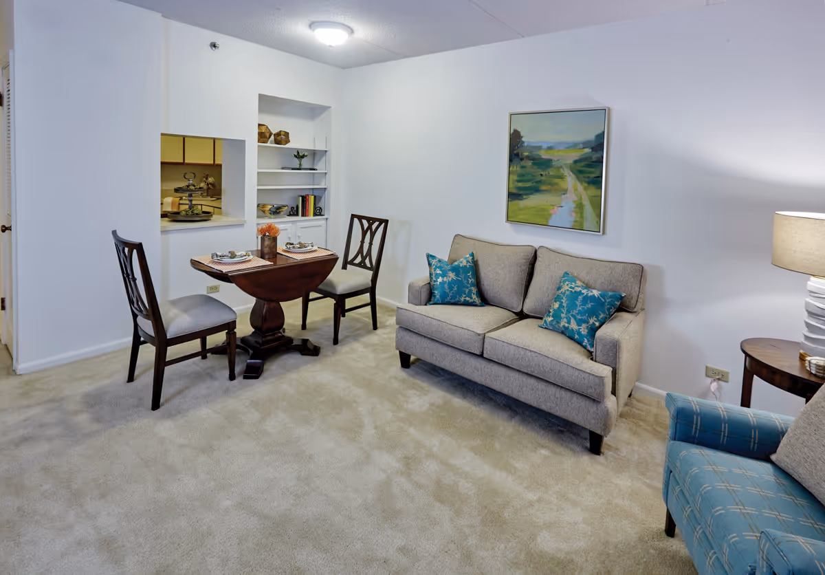 A cozy living area in a senior living facility featuring a small wooden dining table set with two chairs, a gray loveseat with two blue patterned pillows, a blue armchair, a side table with a lamp, and a landscape painting on the wall. There is a pass-through window to the kitchen and built-in shelves with decorative items.
