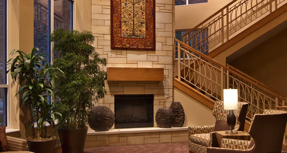A cozy indoor seating area featuring a stone fireplace with a wooden mantle and a decorative hanging above it. Two large potted plants are placed near the fireplace. There are patterned armchairs and a small table with a lit lamp. A staircase with a wooden handrail and decorative metal balusters is visible in the background.