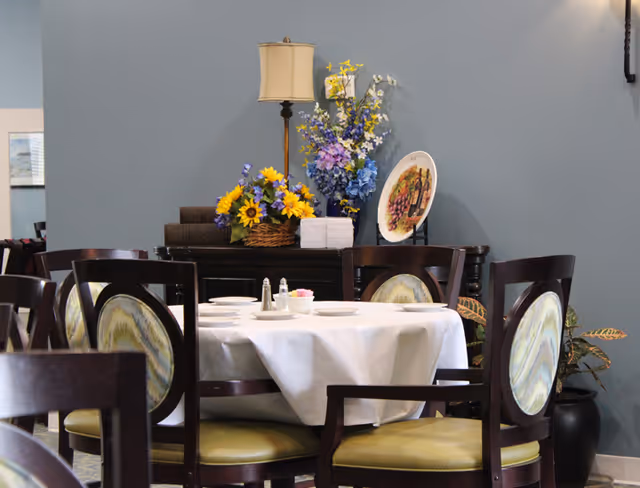 Round dining table with a white tablecloth and place settings surrounded by chairs in a decorated dining area with flowers and a lamp.