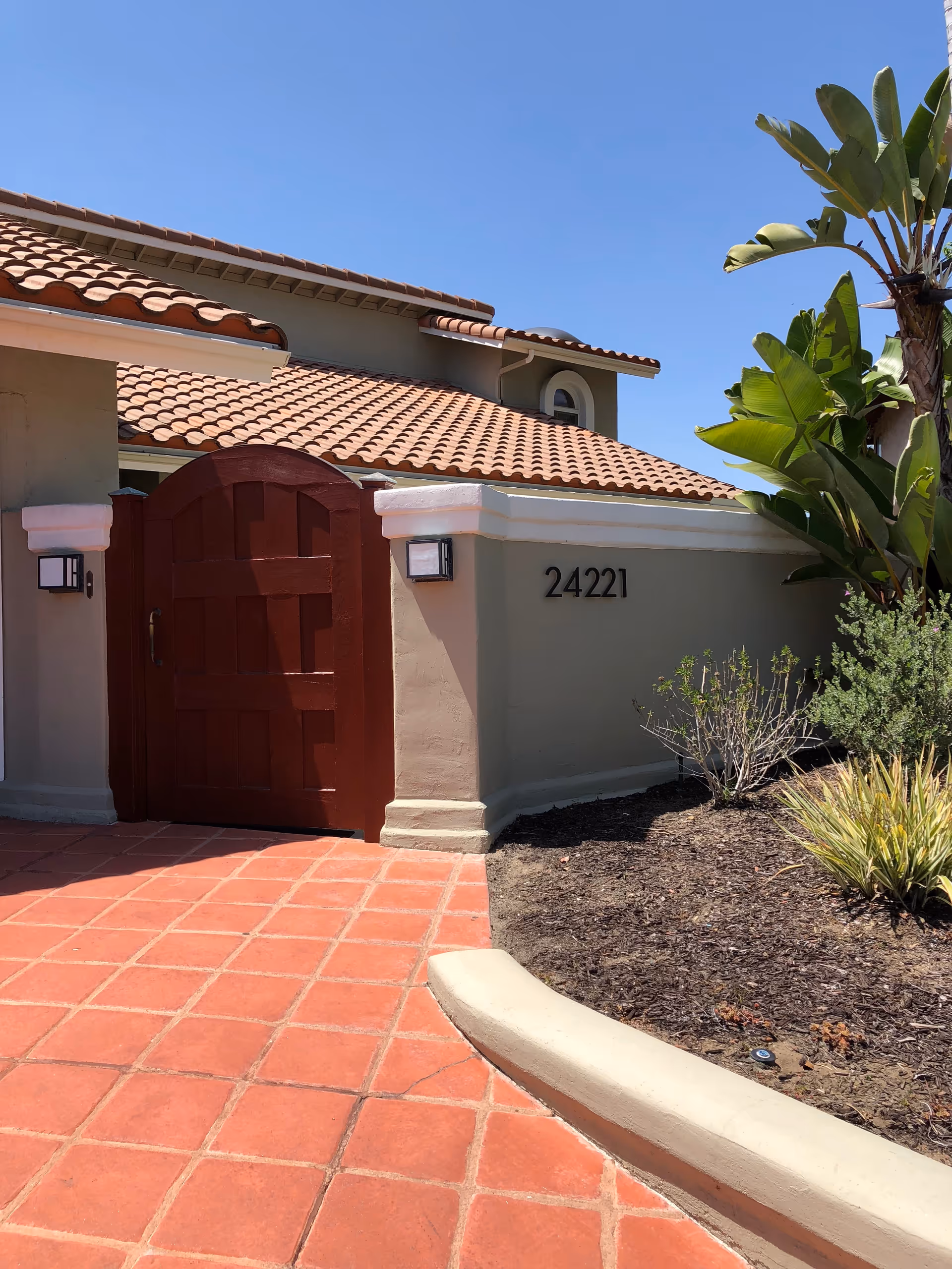Exterior view of a building with a red tiled roof and a red wooden gate set in a beige stucco wall. The building number 24221 is displayed on the wall. There are plants and shrubs to the right of the gate and a clear blue sky above.