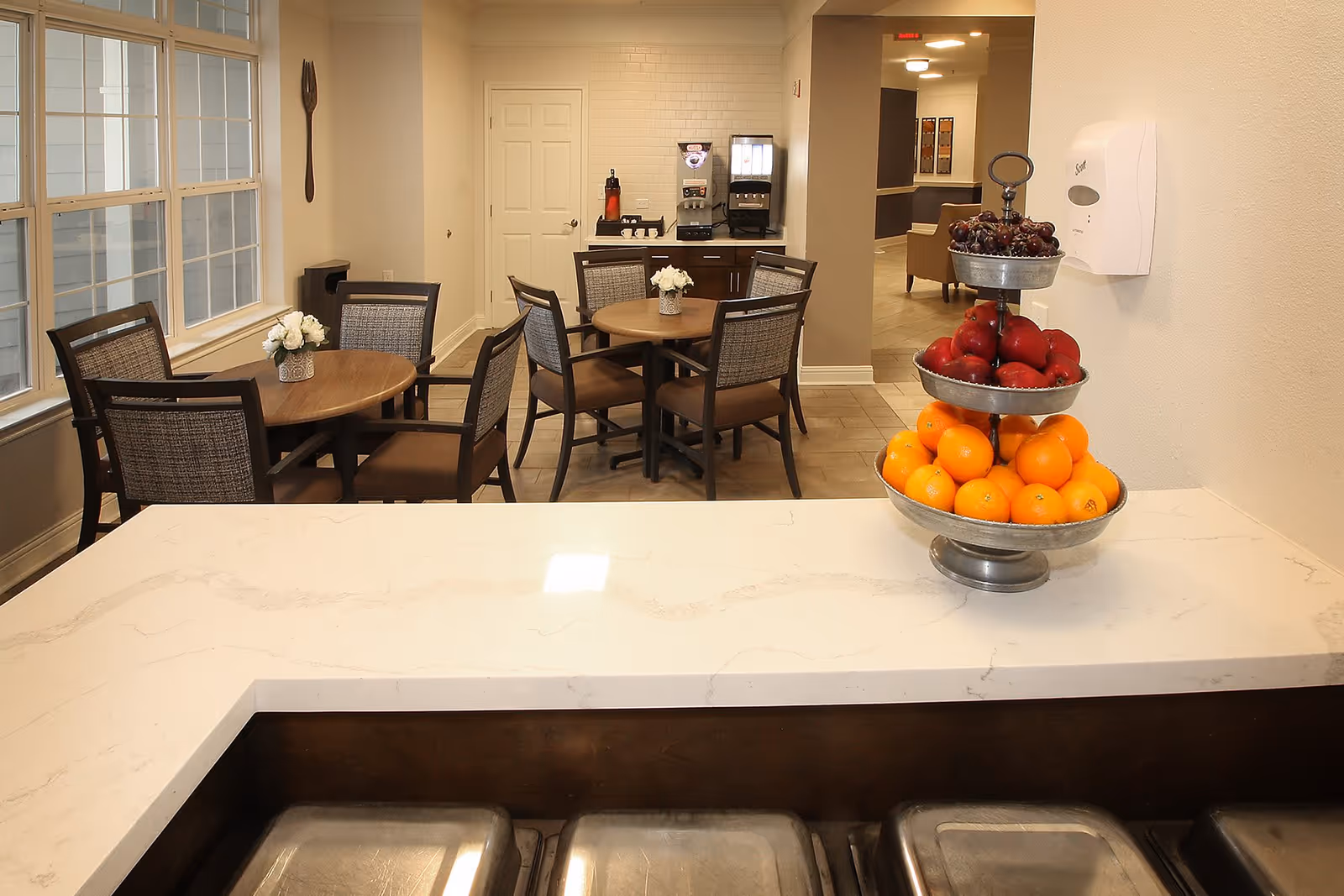Interior view of a senior living facility dining area with round wooden tables and chairs. A three-tiered fruit stand with oranges, red apples, and grapes is placed on a white countertop in the foreground. In the background, there is a beverage station with a coffee machine and a water dispenser, and a hallway leading to another room.