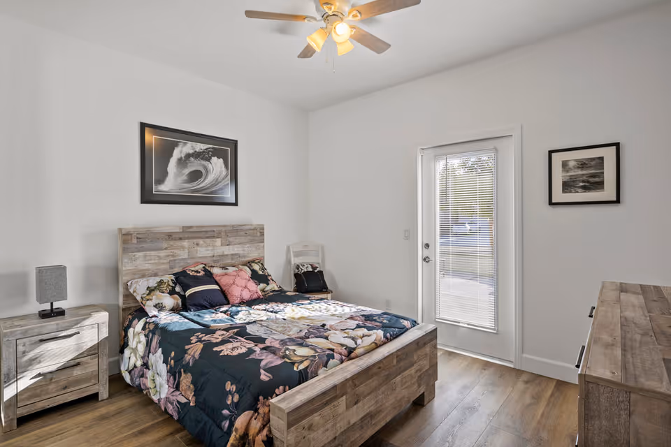 A bright bedroom with a wooden bed frame and matching nightstand and dresser. The bed is made with a floral patterned comforter and several pillows. There is a ceiling fan with lights above, a framed picture of a wave on the wall above the bed, and a door with blinds leading outside. Another framed picture hangs on the adjacent wall.