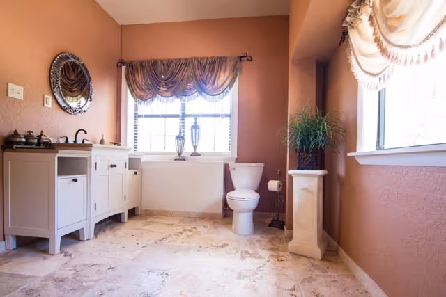A spacious bathroom with beige walls and tiled floor. It features a white vanity with a round decorative mirror above it on the left, a toilet next to a window with sheer curtains in the center, and a tall pedestal with a green plant near another window on the right.