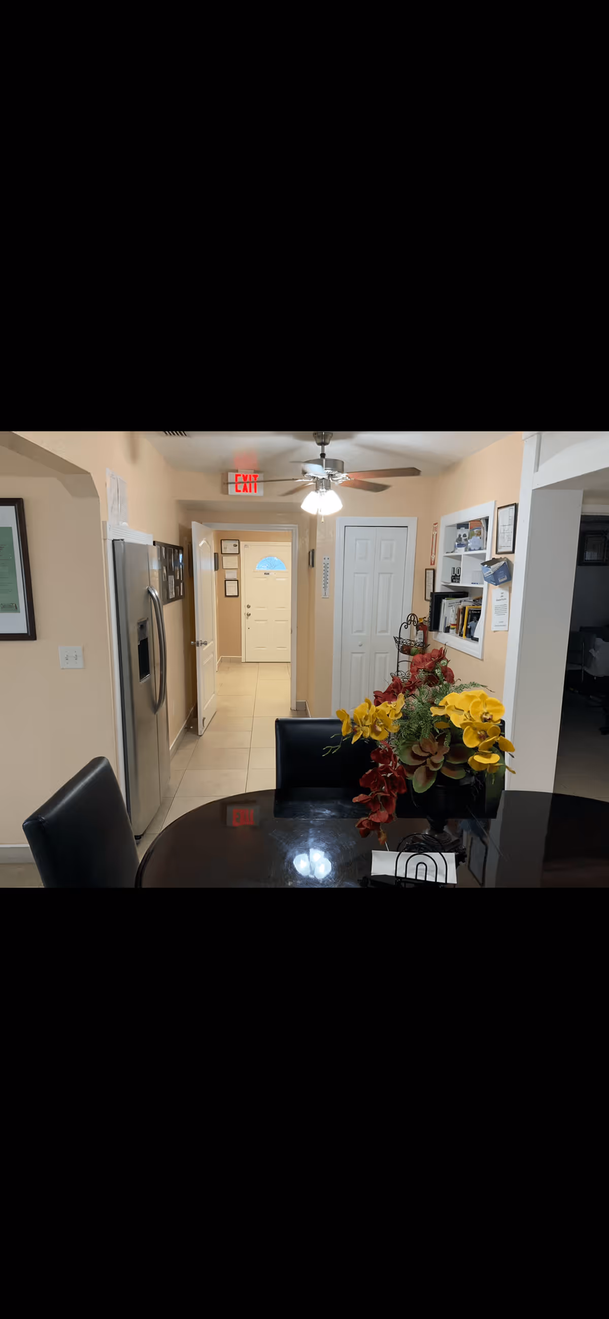 Interior view of a senior living facility dining area with a dark wooden table and black chairs. A floral arrangement with yellow and red flowers is placed on the table. In the background, there is a hallway leading to a white door with certificates and notices on the walls. A ceiling fan with lights is on, and a stainless steel refrigerator is visible on the left side.
