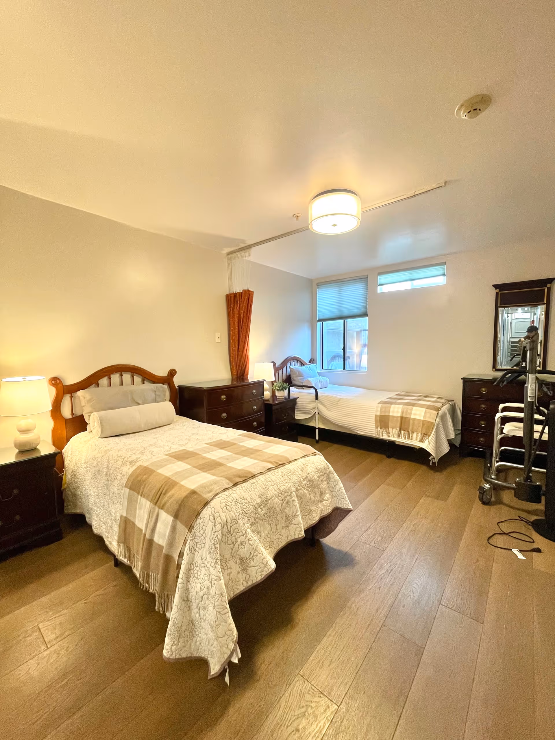 A well-lit bedroom with two single beds, each with a wooden headboard, white bedding, and a beige plaid throw blanket. There are two dark wooden nightstands with lamps, a dresser with a mirror, and a walker near the dresser. The room has wooden flooring and a window with blinds letting in natural light.