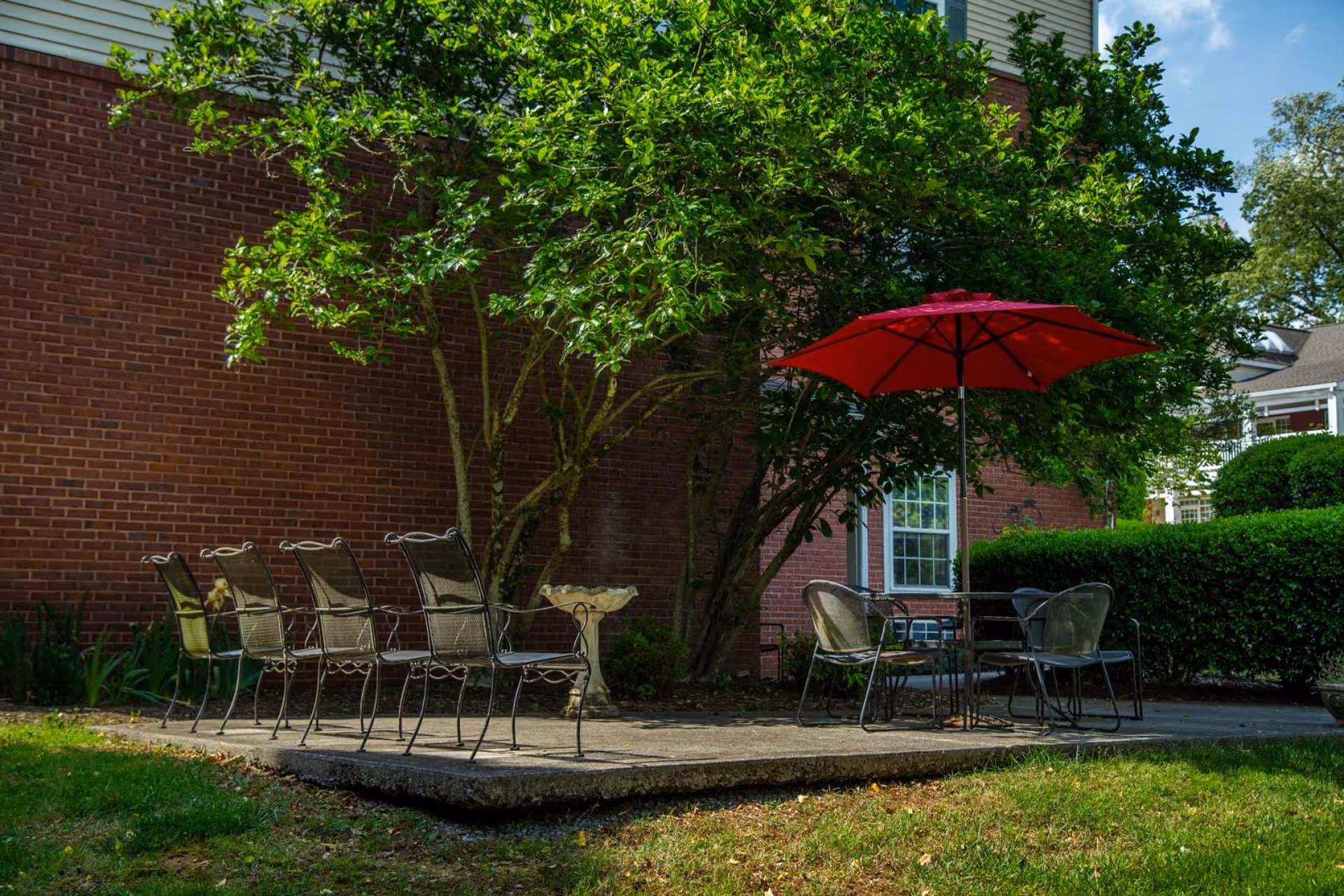 Outdoor patio area with metal chairs arranged in two groups on a concrete slab. One group has six chairs in a row, and the other group has four chairs around a table with a red umbrella. The patio is next to a brick building with a window and surrounded by green bushes and trees.